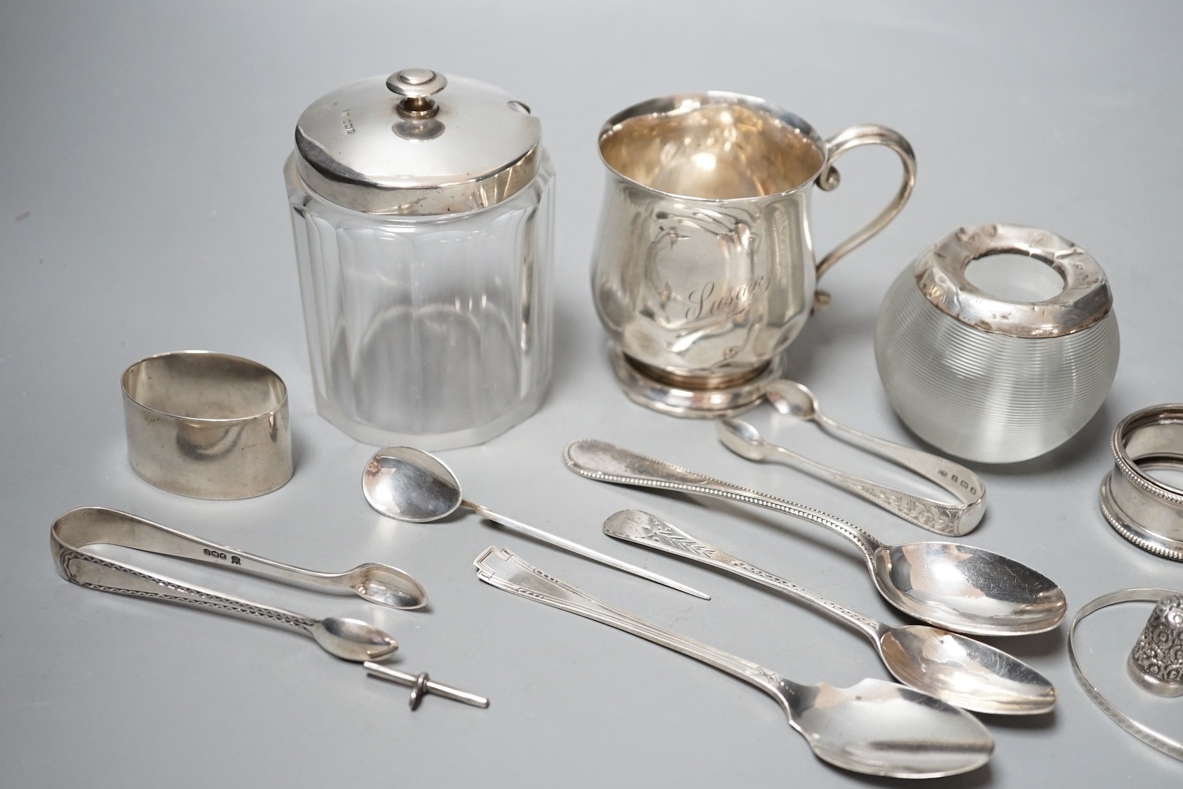 Sundry small silver including a mounted glass match tidy(a.f.), a silver christening mug(a.f.), mounted glass preserve jar, three napkin rings, six items of flatware, thimble T-bar and a sterling bangle.